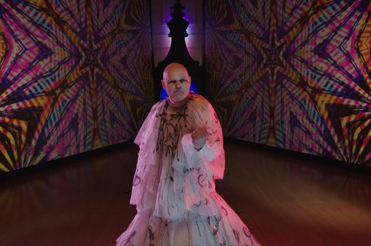 William Corgan în the Beguiled Video in white dress against colorful background.