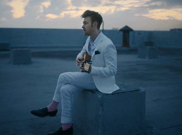 Finneas in a white suit sitting on a rooftop with guitar