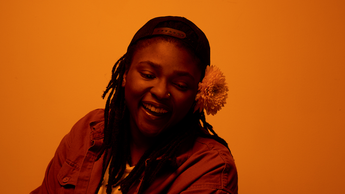 Orange tinted photo of Joy Oladokun smiling wearing baseball hat.