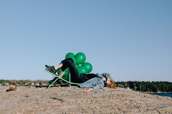 photo of artist laying on beach in collapsed chair with green balloons.