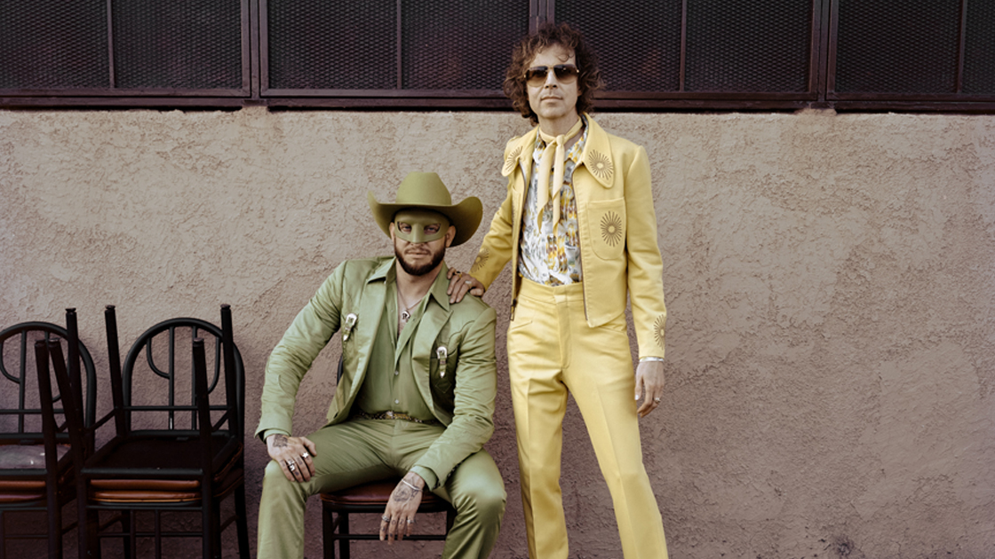 Two men wearing western wear outside.