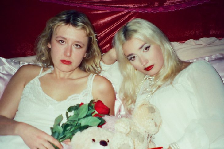 Two women dressed in white laying on pink satin bed.