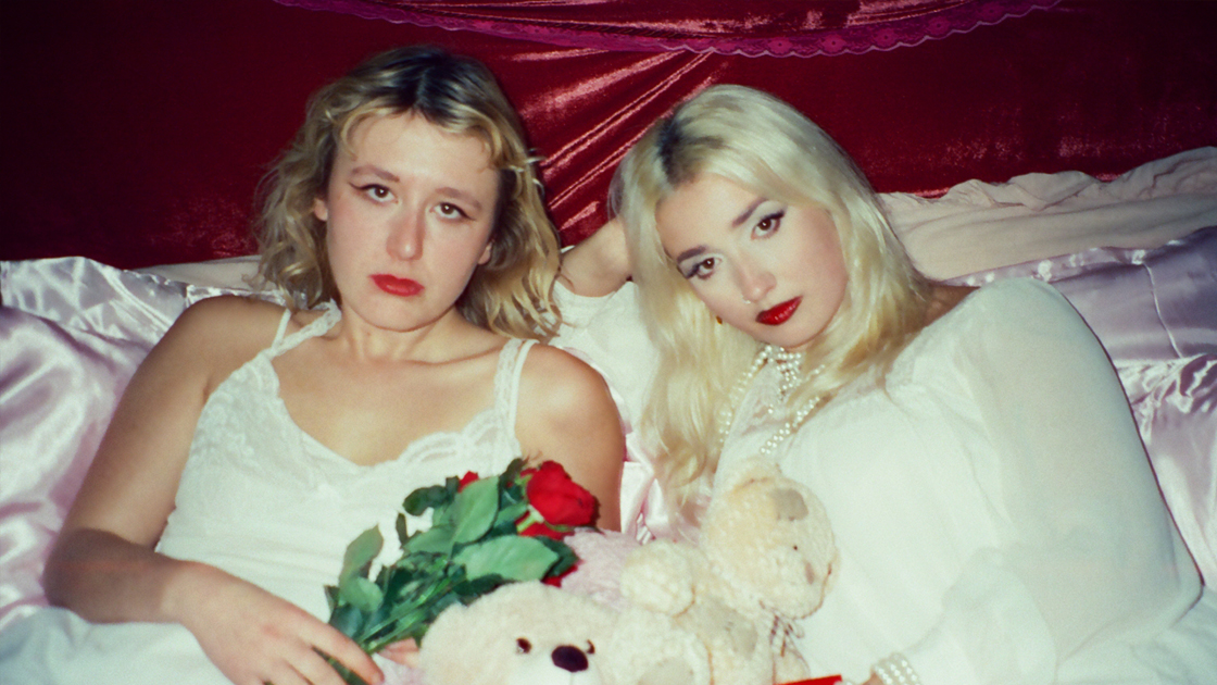 Two women dressed in white laying on pink satin bed.