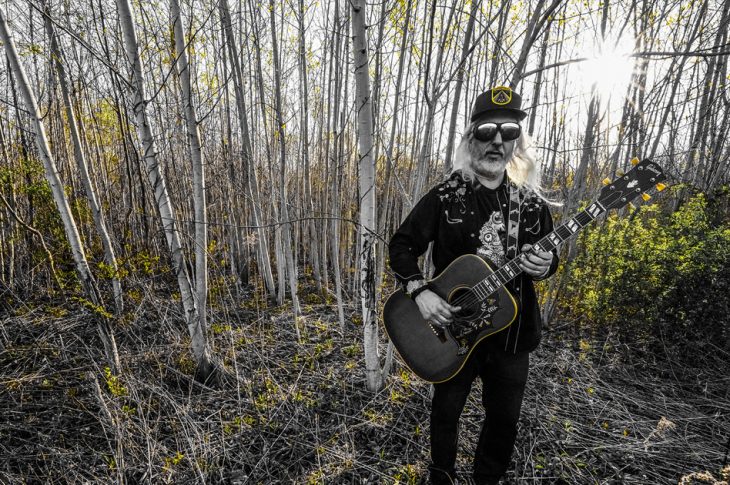 Man with acoustic guitar standing in woods.