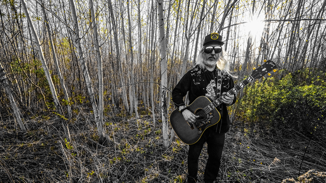 Man with acoustic guitar standing in woods.