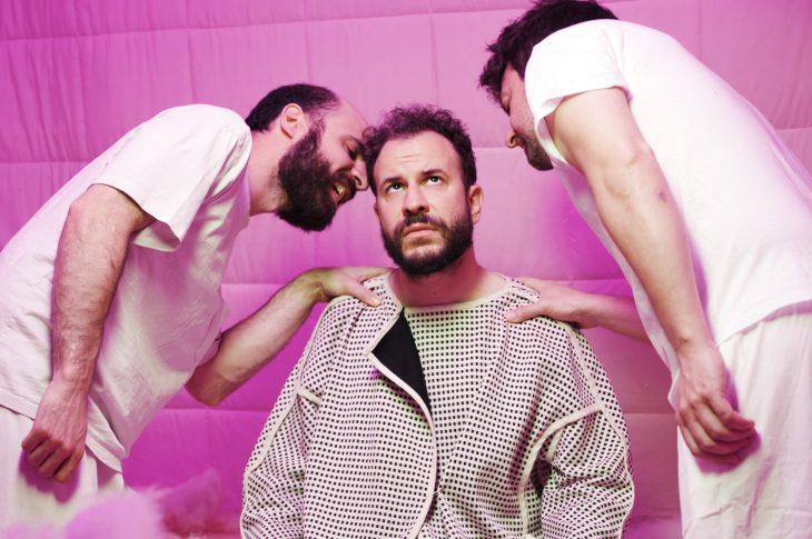 three people in a pink room. One sits in a hospital gown while two others whisper in the their ear.