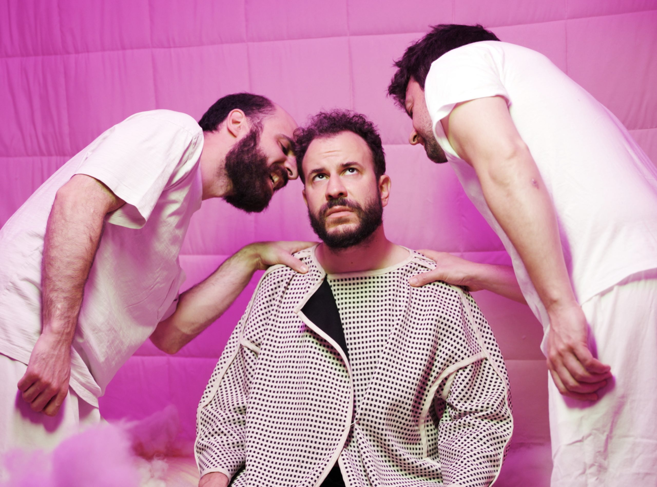three people in a pink room. One sits in a hospital gown while two others whisper in the their ear.