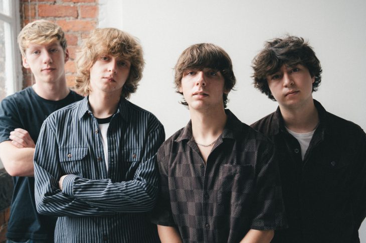 Four men photographed in room with one white wall and one brick wall.