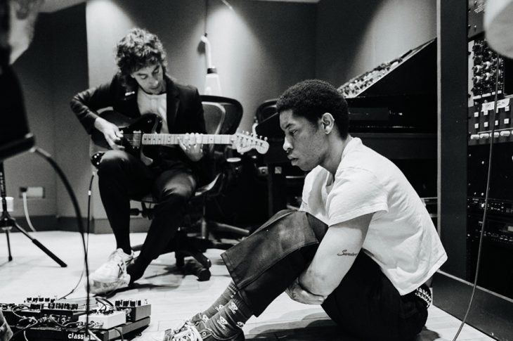 Two men working in the studio. 1 with a guitar and 1 sitting on a floor.
