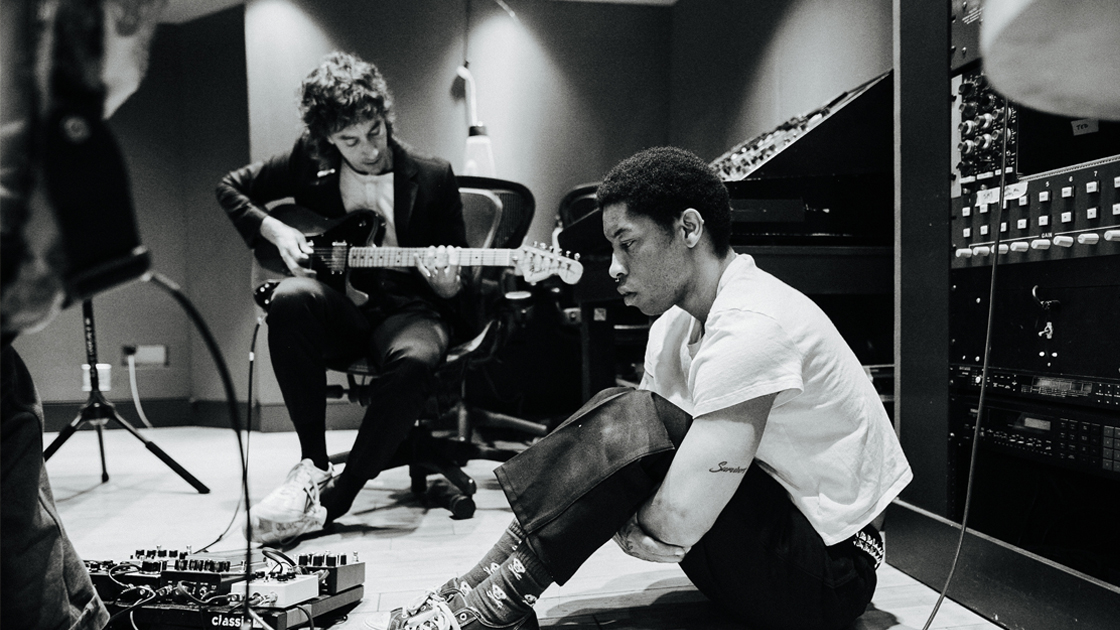Two men working in the studio. 1 with a guitar and 1 sitting on a floor.