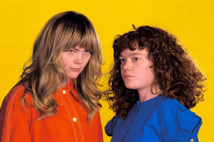 Two people in orange and blue shirts photographed against yellow background.