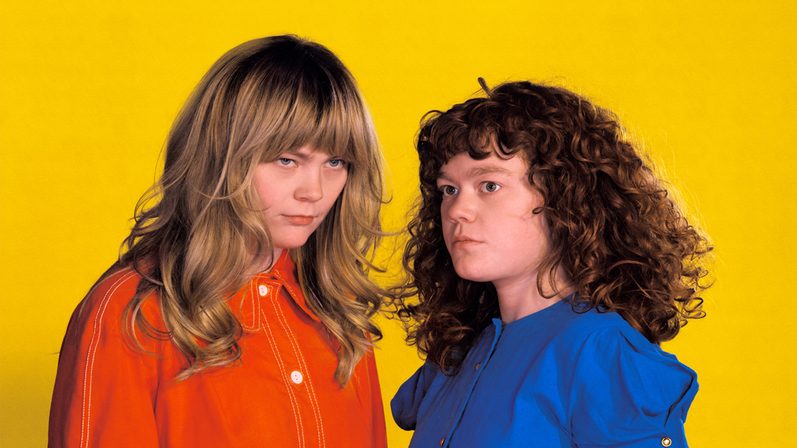 Two people in orange and blue shirts photographed against yellow background.